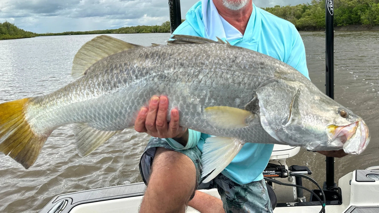 An angler in Tackle World gear holds a large barramundi on his boat with Power-Pole anchors amid mangrove-lined creeks under an overcast sky.