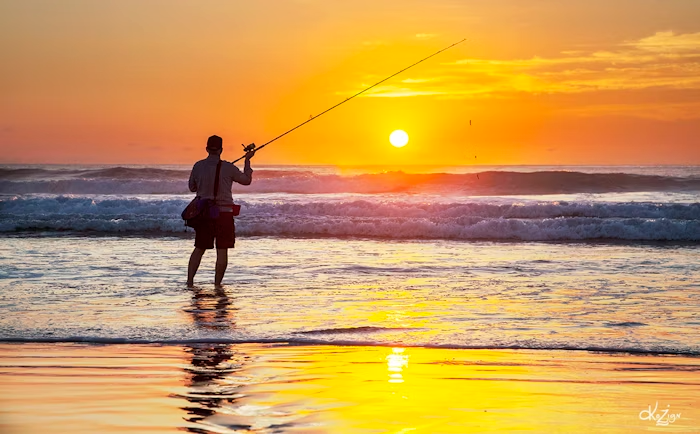 A beginner learning beach fishing techniques, standing on a sandy shore with a fishing rod, casting into the waves under a clear sky