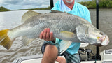 An angler in Tackle World gear holds a large barramundi on his boat with Power-Pole anchors amid mangrove-lined creeks under an overcast sky.