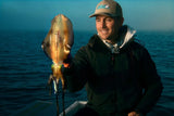 An angler wearing a Tackle World cap holds a vibrant, freshly caught squid on a boat over calm blue ocean waters.